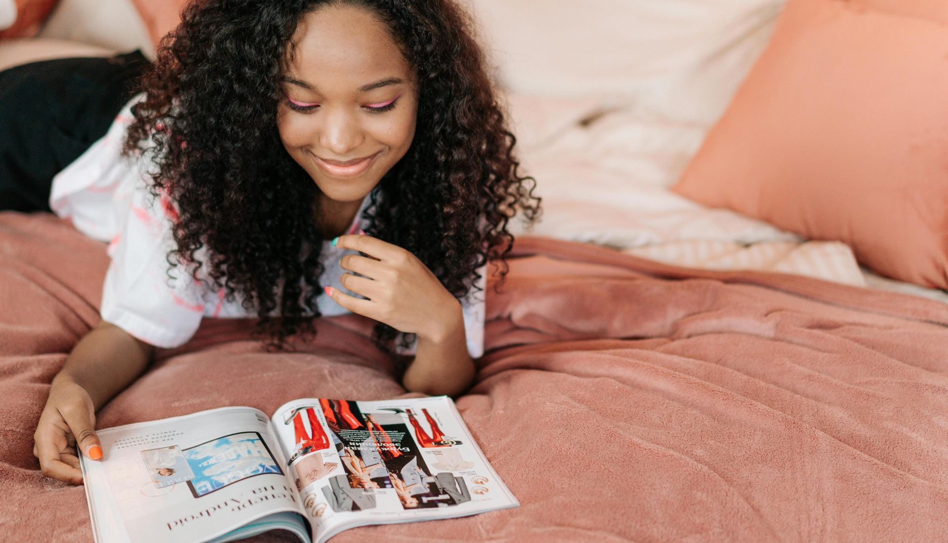 Smiling teenager with curly hair reading a magazine on a bed with peach-colored pillows.